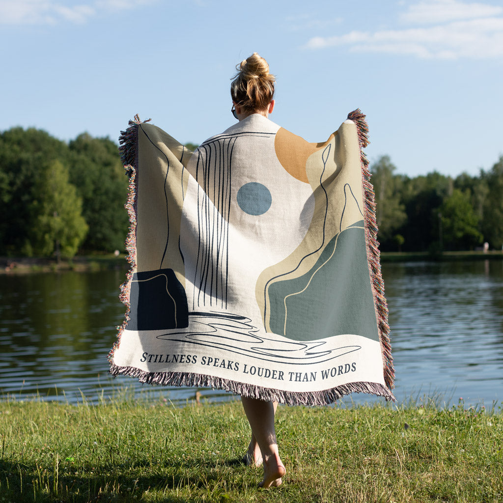 Person holding a decorative blanket with abstract patterns and text by a lake.