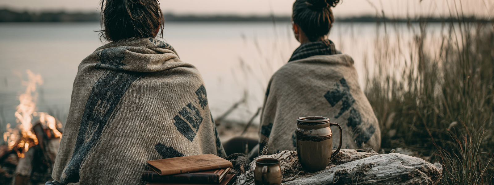 Two people wrapped in blankets sitting by a campfire by a lake.